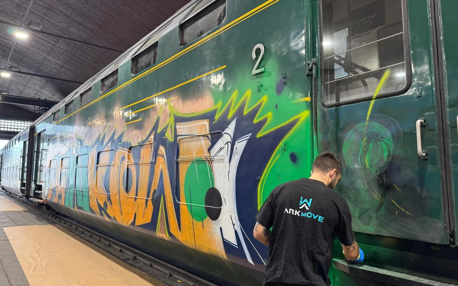 Bok zeleného patrového vozu ČSD pokrytý velkoplošným graffiti, pracovník ARK move ho v depu čistí